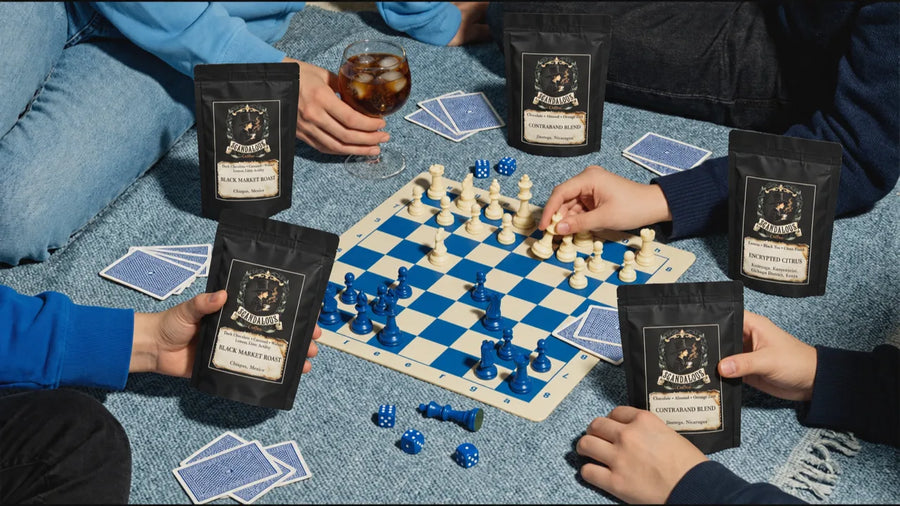Five Scandalous Coffee bags around a blue chessboard on a picnic blanket with cards, dice and iced coffee.