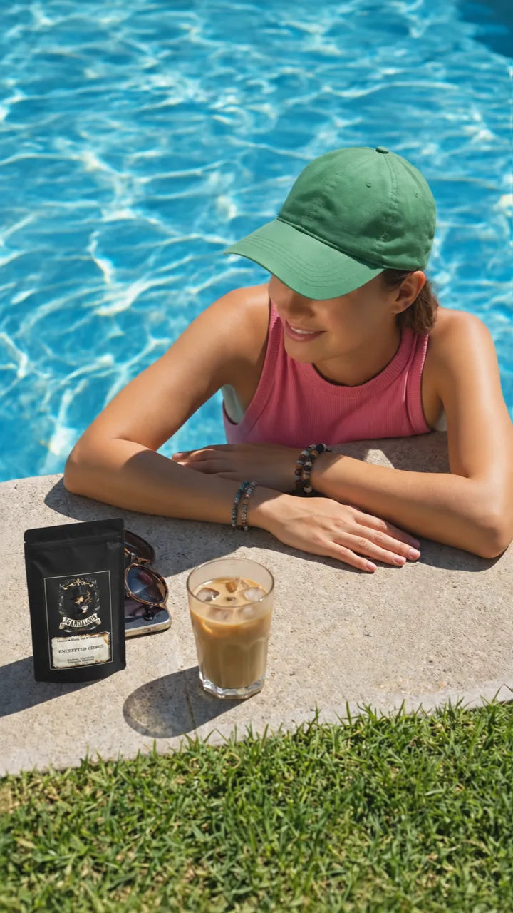 A woman in a green cap relaxes by a blue pool beside iced coffee and an Encrypted Citrus bag.