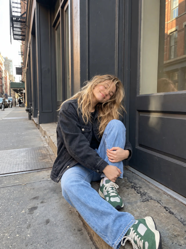 A woman in a dark denim jacket and blue jeans sits on a city sidewalk beside a charcoal storefront.