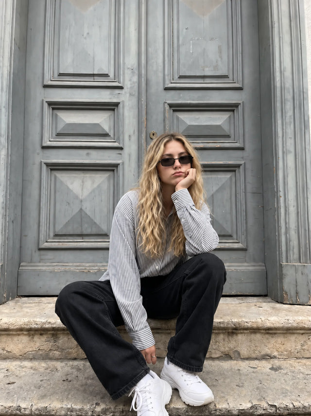 A woman in dark sunglasses, a striped shirt and black jeans sits on stone steps before gray paneled doors.